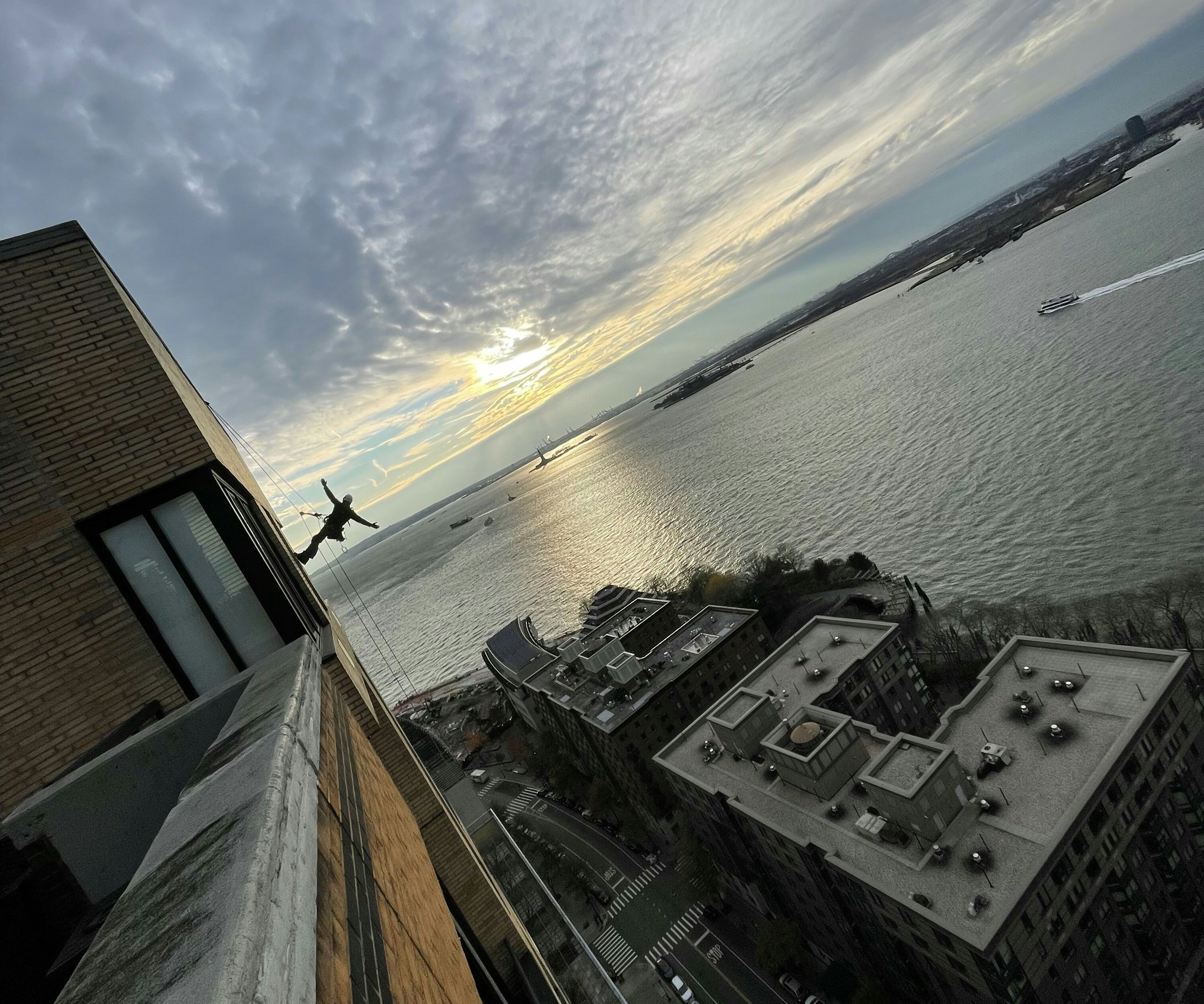 <p>A Rope Access Inspector for FISP in full safety gear is suspended by ropes on the side of a tall building, silhouetted against a dramatic sunset over a wide river.</p>